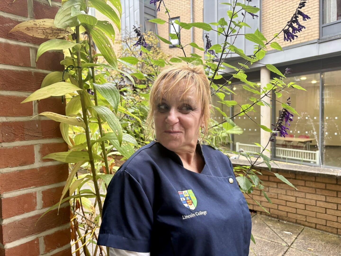 Mary O’Boyle, a Lincoln College scout, standing in a courtyard beside a brick pillar and flowering plants, with College buildings visible in the background.