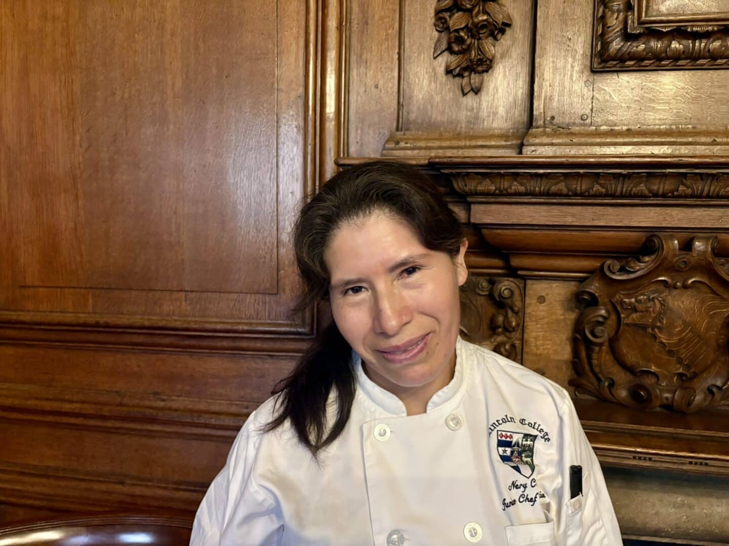 Nery Cucho Najarro, Junior Chef de Partie at Lincoln College, seated in front of an ornate wooden fireplace with carved detailing, wearing a white chef’s jacket embroidered with her name and the College crest.