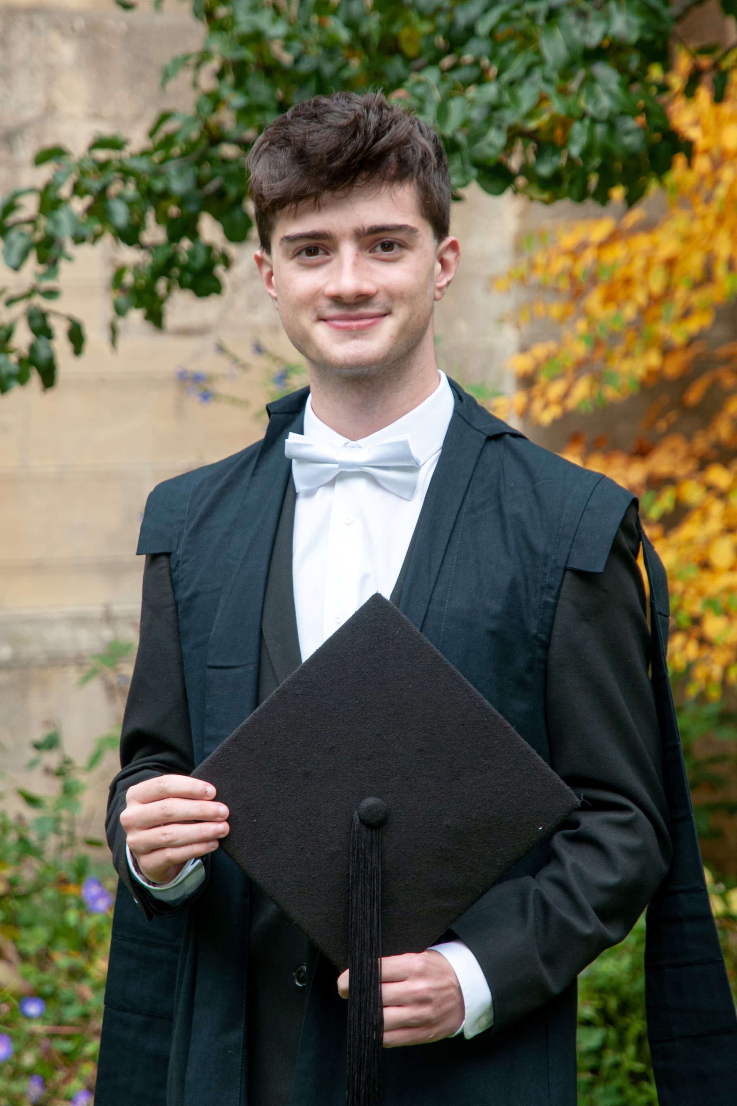 Headshot of Ben Wheadon, a young man in subfusc gown with a white bowtie, holding his mortarboard.