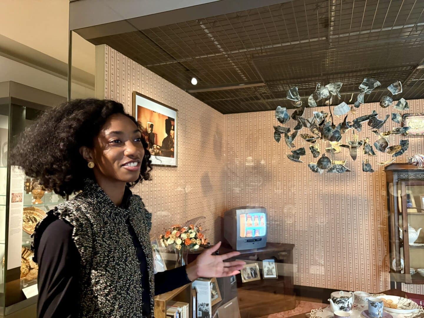 Olivia Holder stands in a museum gallery, gesturing toward a glass display case that recreates a domestic room scene. Inside the case are patterned wallpaper, a small television, framed photos, a vase of flowers, and a tea set on a table. Suspended above is an art installation of fragmented ceramic pieces hanging from the ceiling. She looks toward the display, appearing to explain or present the exhibit.