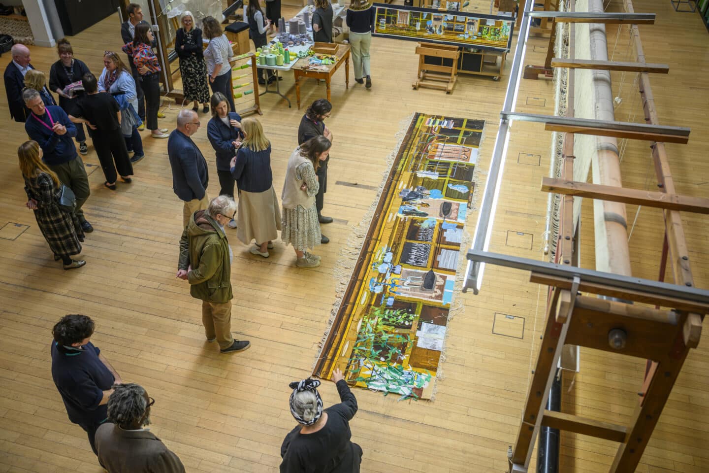 A crowd gathered around the tapestry laid flat on the wooden floor at Dovecot Studios, examining its intricate details and textures.