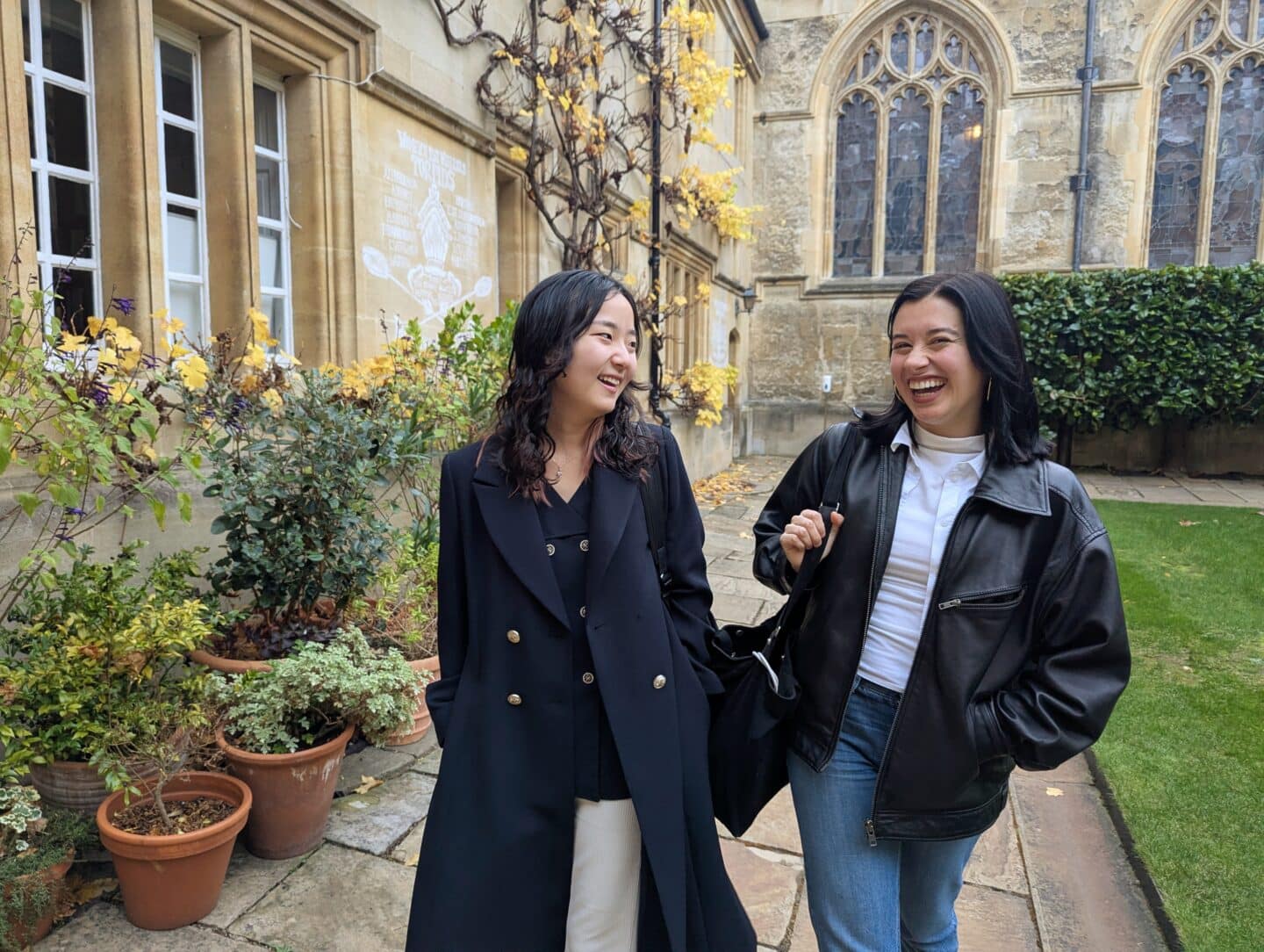 Monica Ruiz and Jaeah Kim walk together through Chapel quad, smiling and chatting. Jaeah wears a long black coat, and Monica wears a black leather jacket with jeans. Behind them are potted plants and the Chapel with arched windows and autumn leaves.