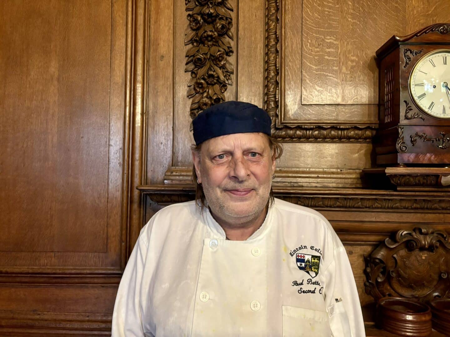 Paul Butterfield, Second Chef at Lincoln College, standing in front of an ornate wooden fireplace with carved detailing and a clock above, wearing a white chef’s jacket and navy cap.