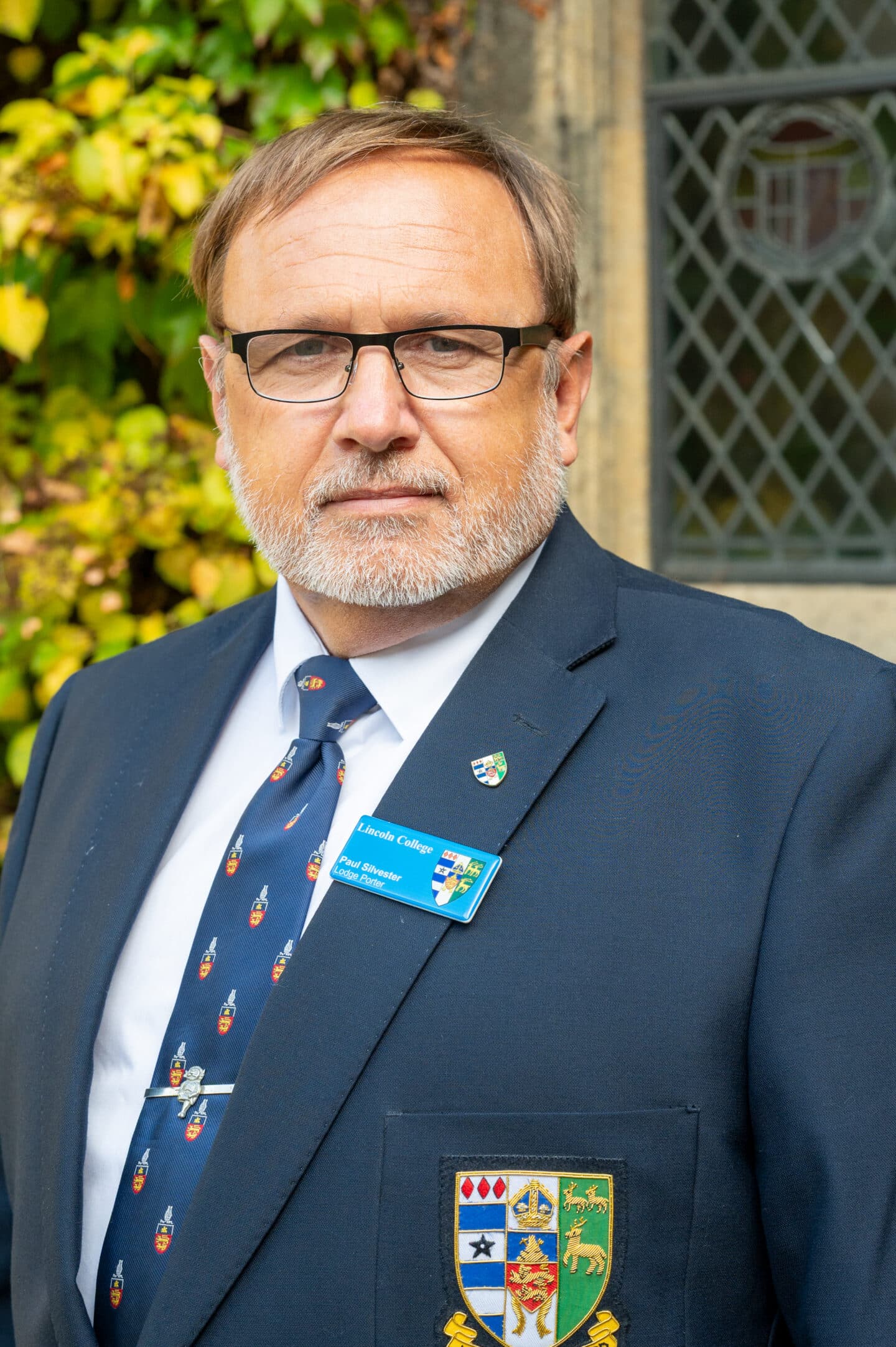 Paul Silvester standing in Front Quad at Lincoln College, Oxford, wearing a suit and College tie, with ivy and a stone window behind.