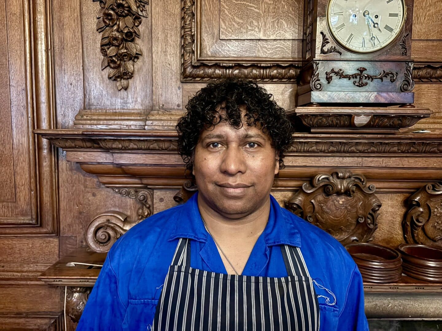 Pedro Gonzaga, a Lincoln College kitchen porter, standing in front of an ornate wooden fireplace with carved detailing and a clock above.