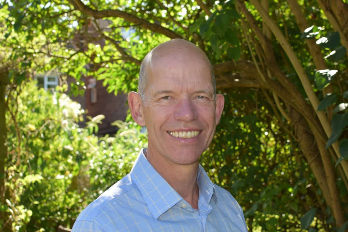Peter Dawson smiling outdoors in a sunlit garden. He is wearing a light blue checked shirt, with leafy trees and a brick building softly blurred in the background.