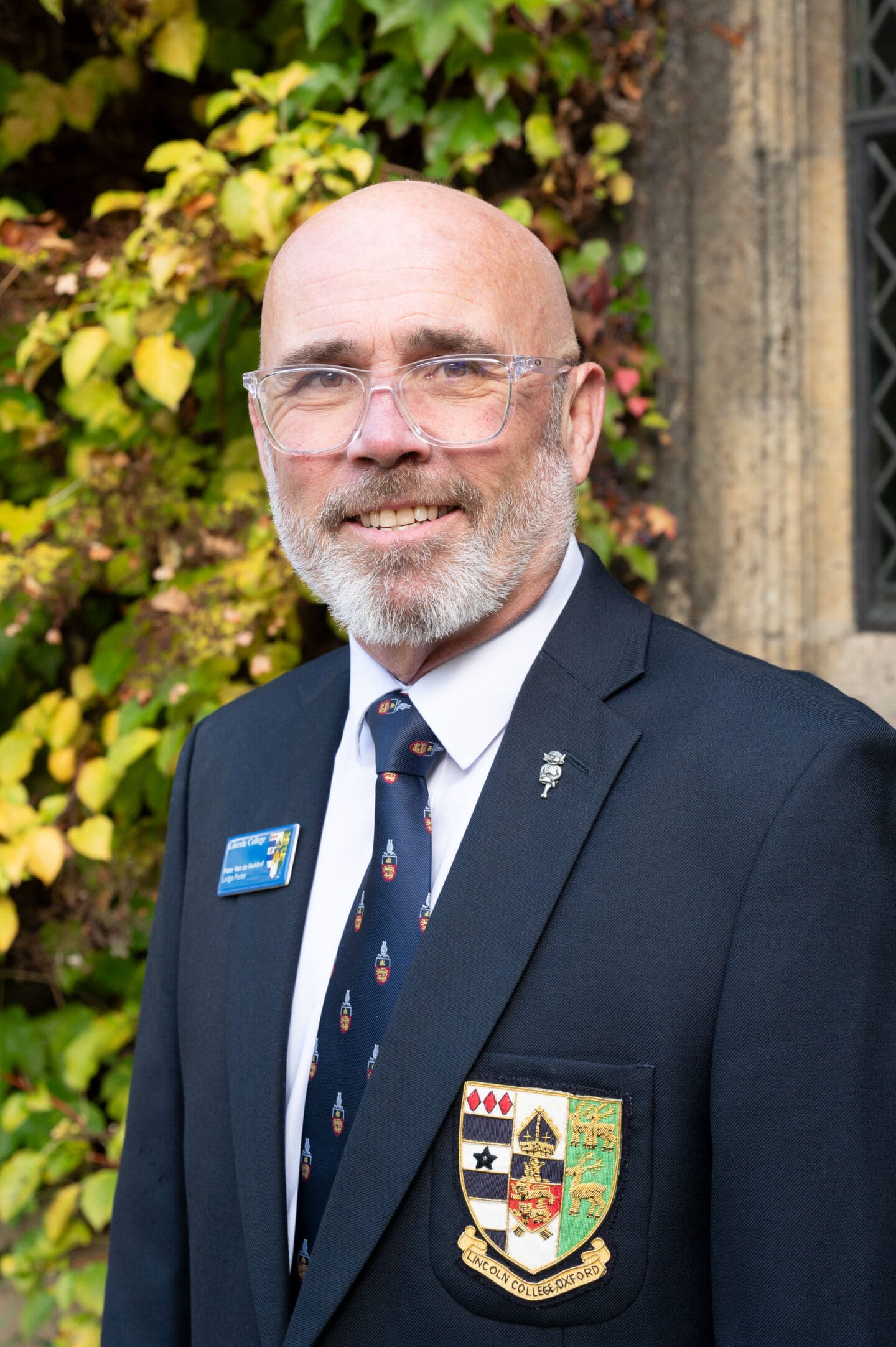 Peter van de Kerkhof standing in Front Quad at Lincoln College, Oxford, wearing a suit and College tie, smiling in front of ivy-covered stonework.