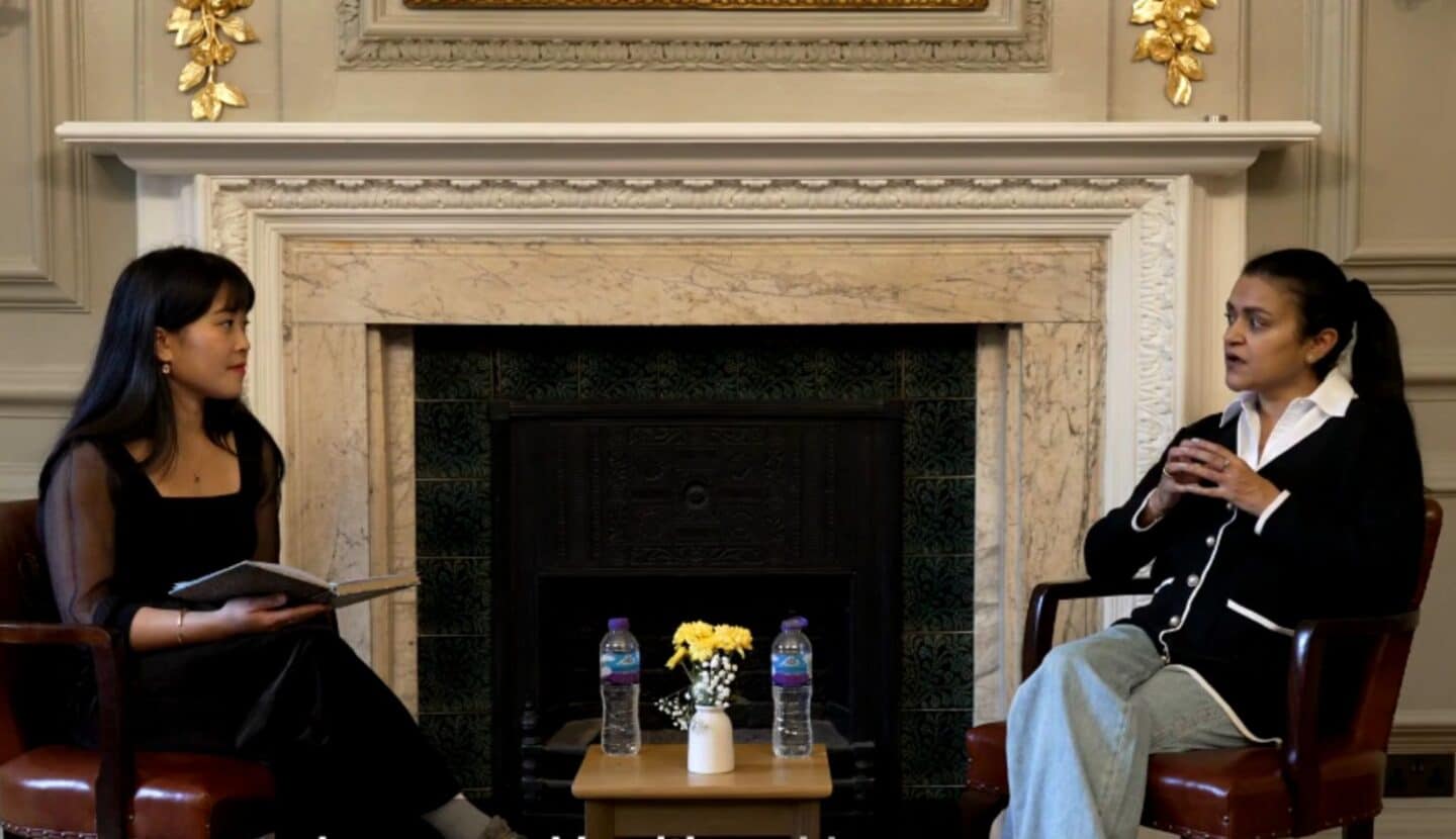 Two women seated in armchairs facing each other in an ornate college room, in conversation in front of a fireplace. Micon Garvilles sits on the left holding notes, listening attentively, while Dr Tara Swart sits on the right speaking and gesturing. A small table between them holds water bottles and flowers, indicating a recorded talk or interview.