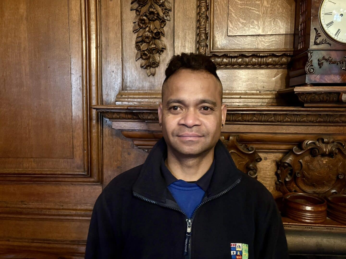 Simon Massey, a Lincoln College scout, seated in front of an ornate wooden fireplace with carved detailing, wearing a fleece with the College crest.