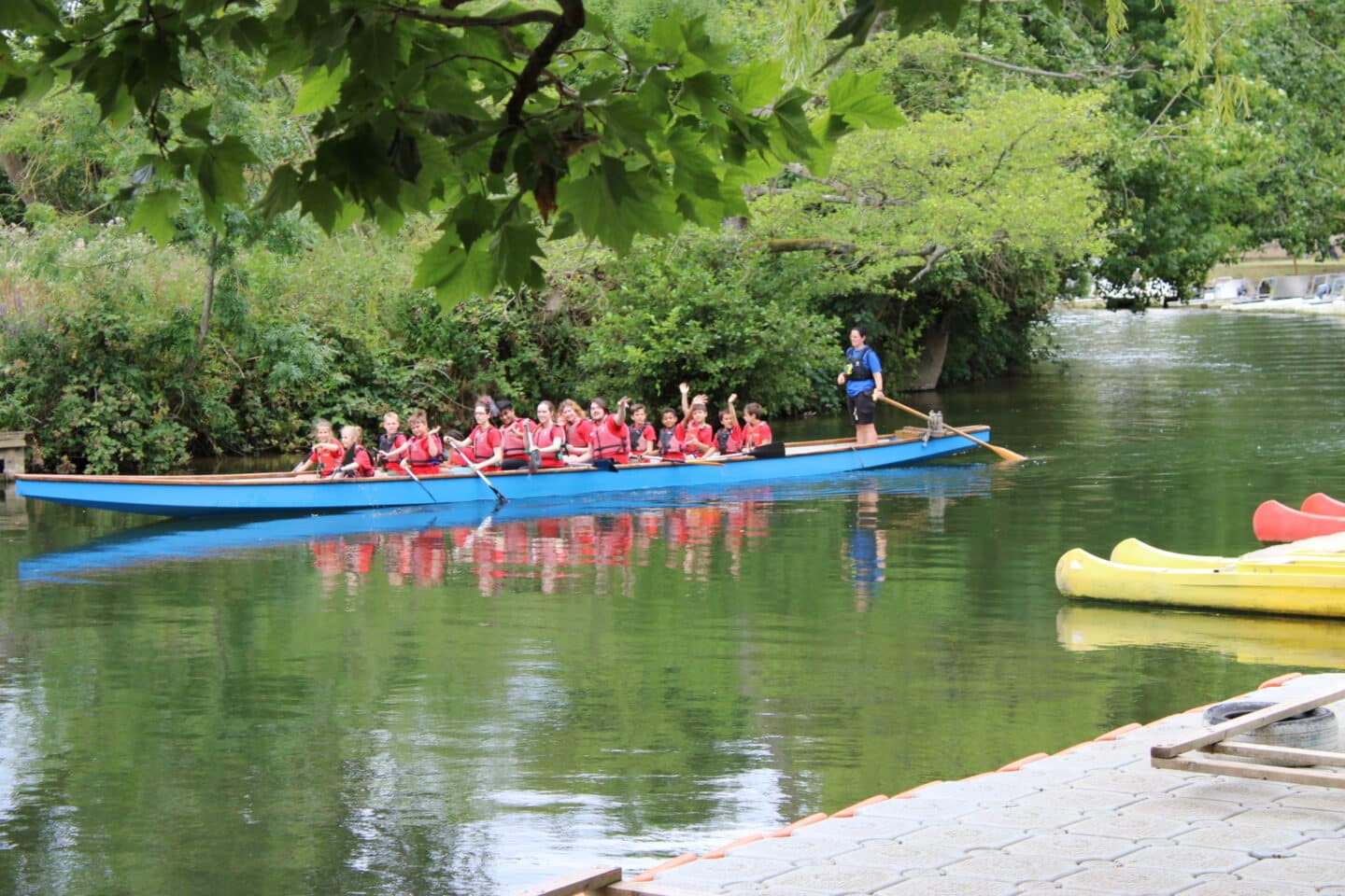 A group of children in red t-shirts and lifejackets rowing along a river in a dragon-boat, with a woman dressed in blue standing at the back of the boat with a large oar.