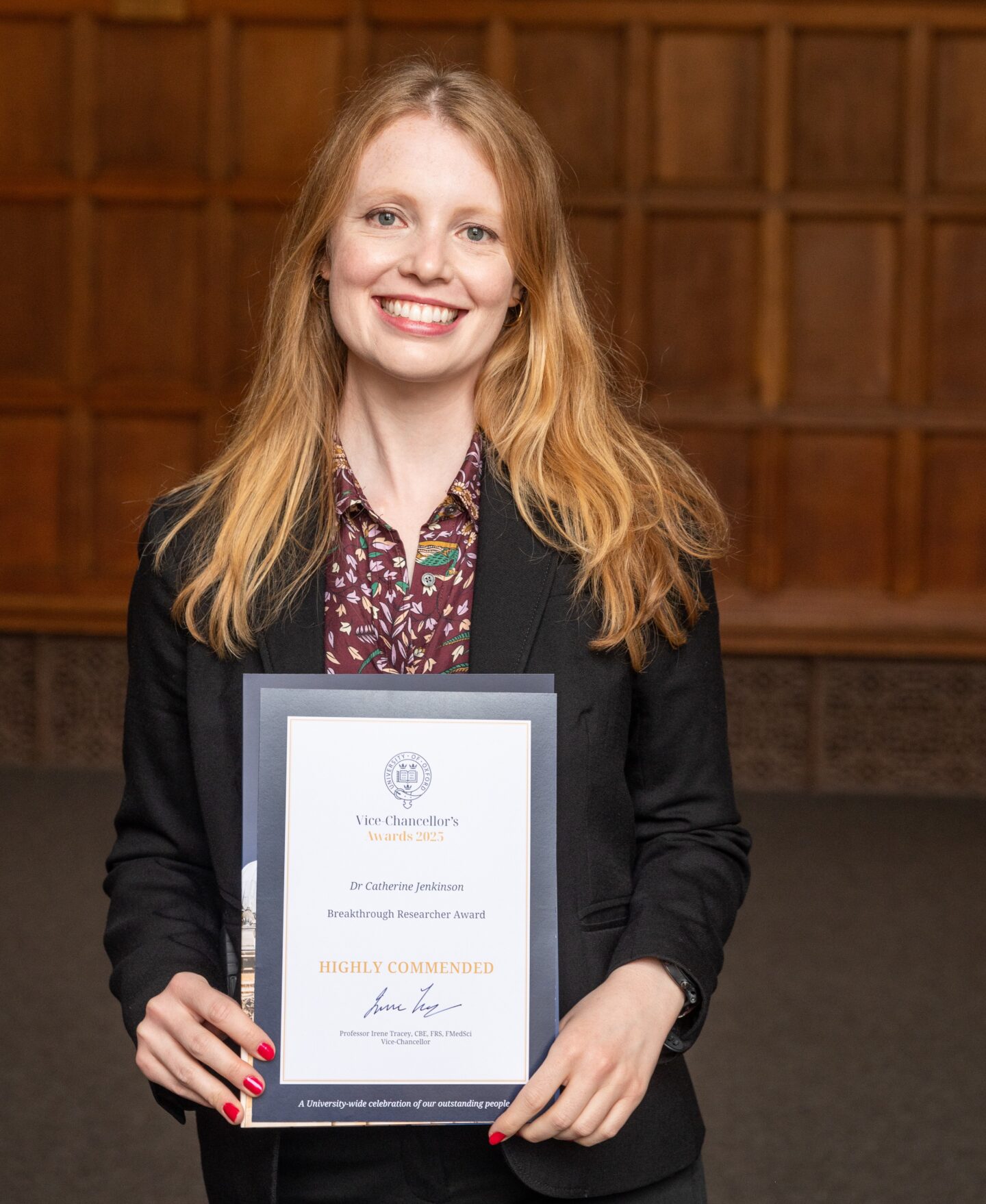 Catherine Jenkinson in black formal jacket and patterned shirt smiling and holding the VC award certificate in front of her