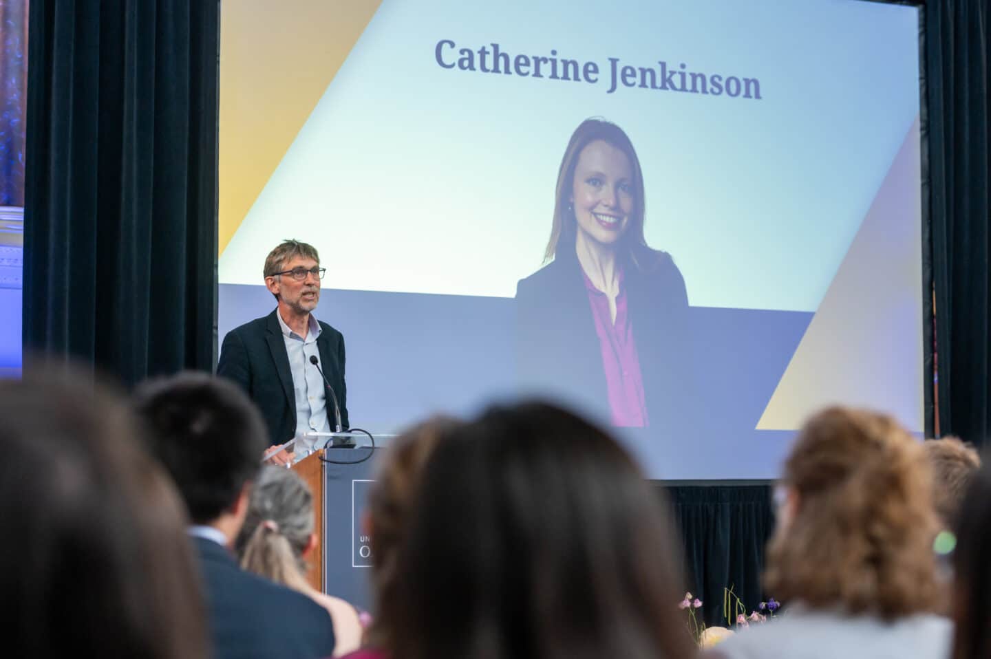 A man stands at a lectern speaking to an audience at the VC awards ceremony. Behind him is a large screen displaying a professional headshot of a smiling Catherine Jenkinson with her name in bold text above her head.