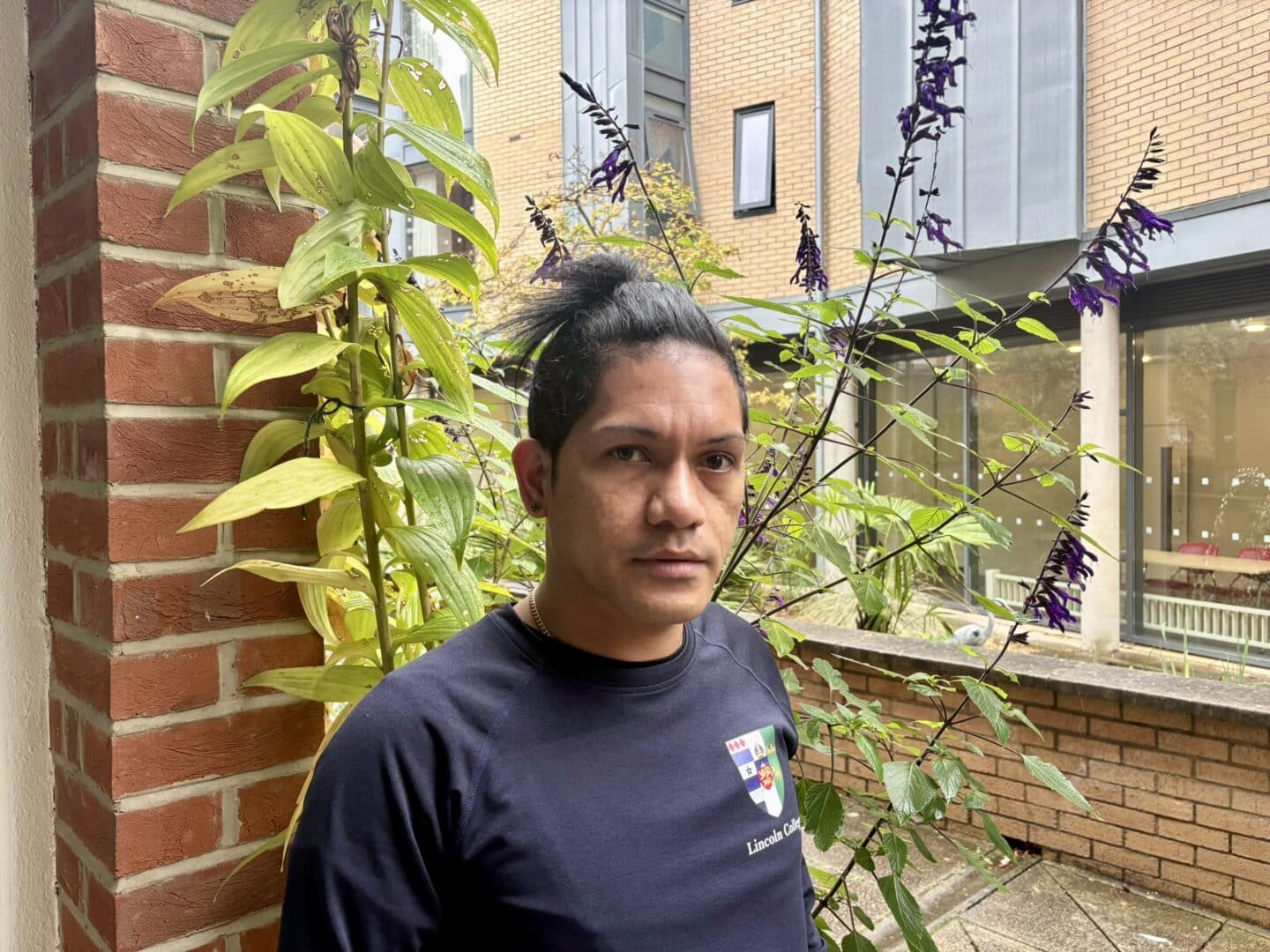Zeca Borges Da Silva, a Lincoln College scout, standing in a courtyard beside a brick pillar and flowering plants, with College buildings visible in the background.