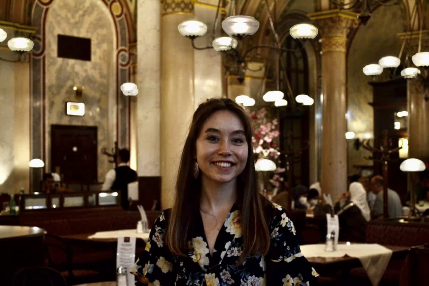 Headshot of Beth Molyneux, a young woman in a floral top, a restaurant in the background