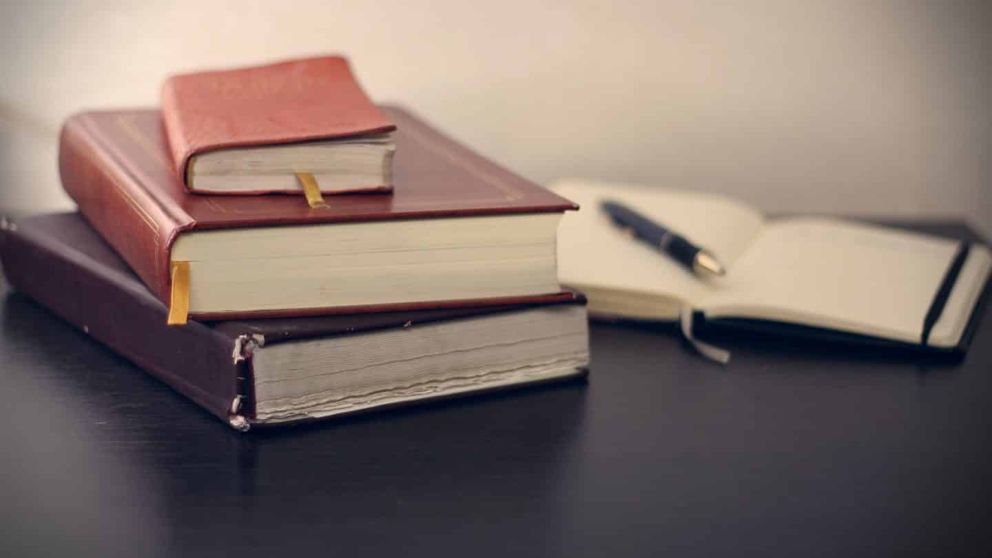 Three closed books on a table with a pen on another open book.