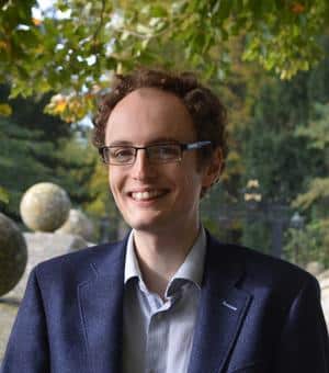 Portrait of a smiling Fred Smith in glasses and a navy blazer, standing in a leafy area.