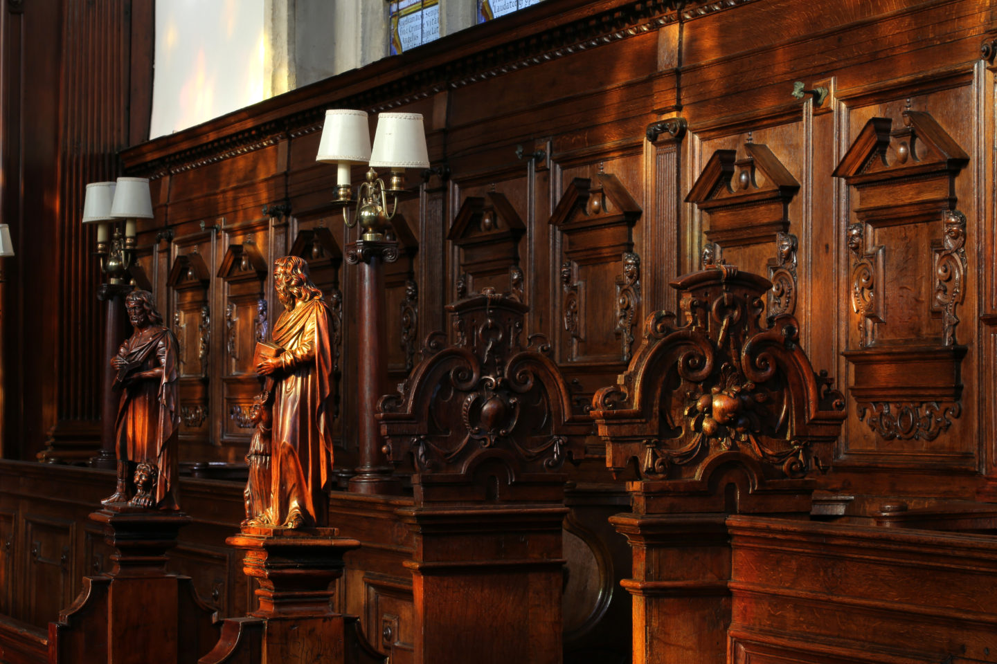 Image of pews in the Lincoln College Chapel, with wooden panelling, wooden benches, two carved wooden figures, and some lamps