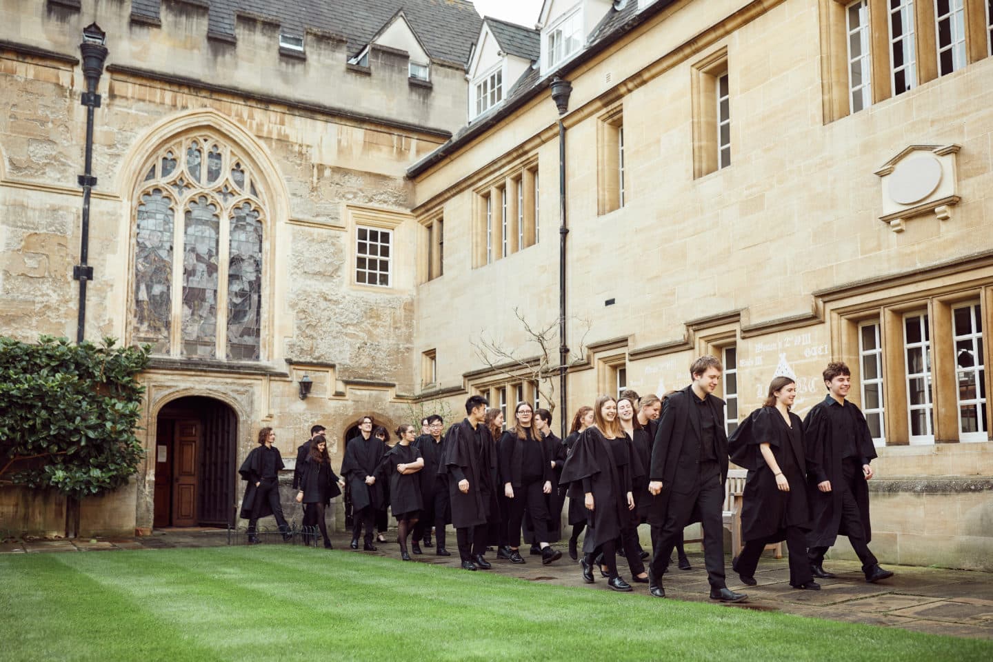 A large group of students dressed in black with gowns walking out of the Chapel into the quad