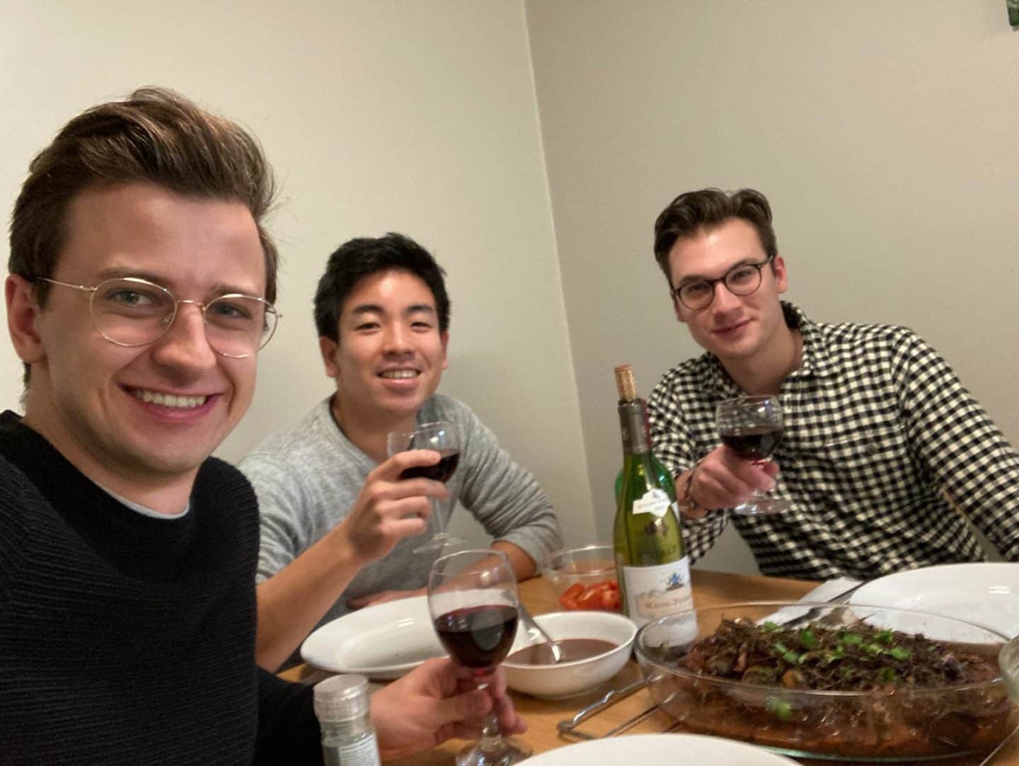 Three young men sitting at a table with food and a bottle, each holding up a glass of red wine