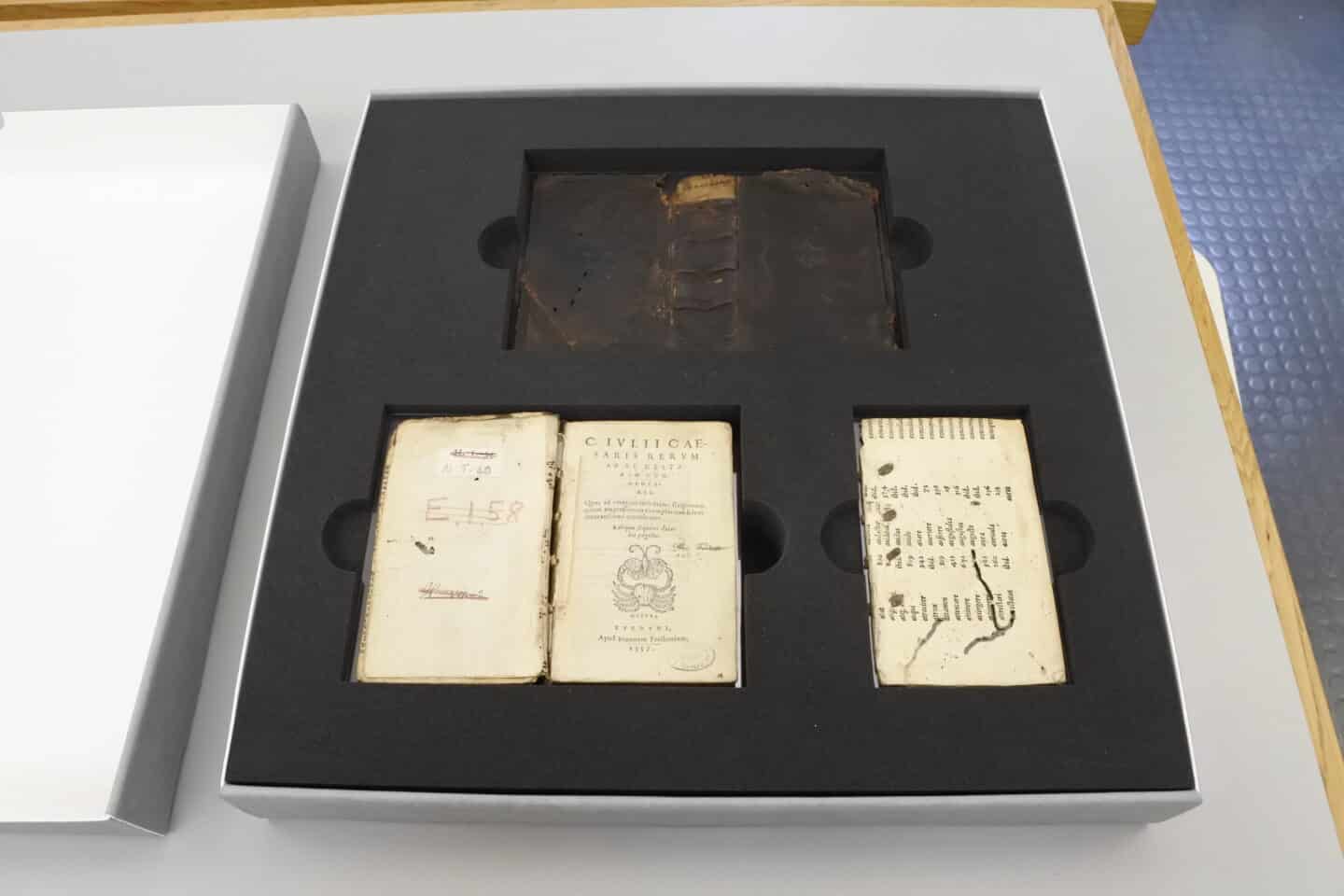 Three sections of a disbound book, including its damaged leather cover and some pages are laid out in a custom-made box with soft protective material