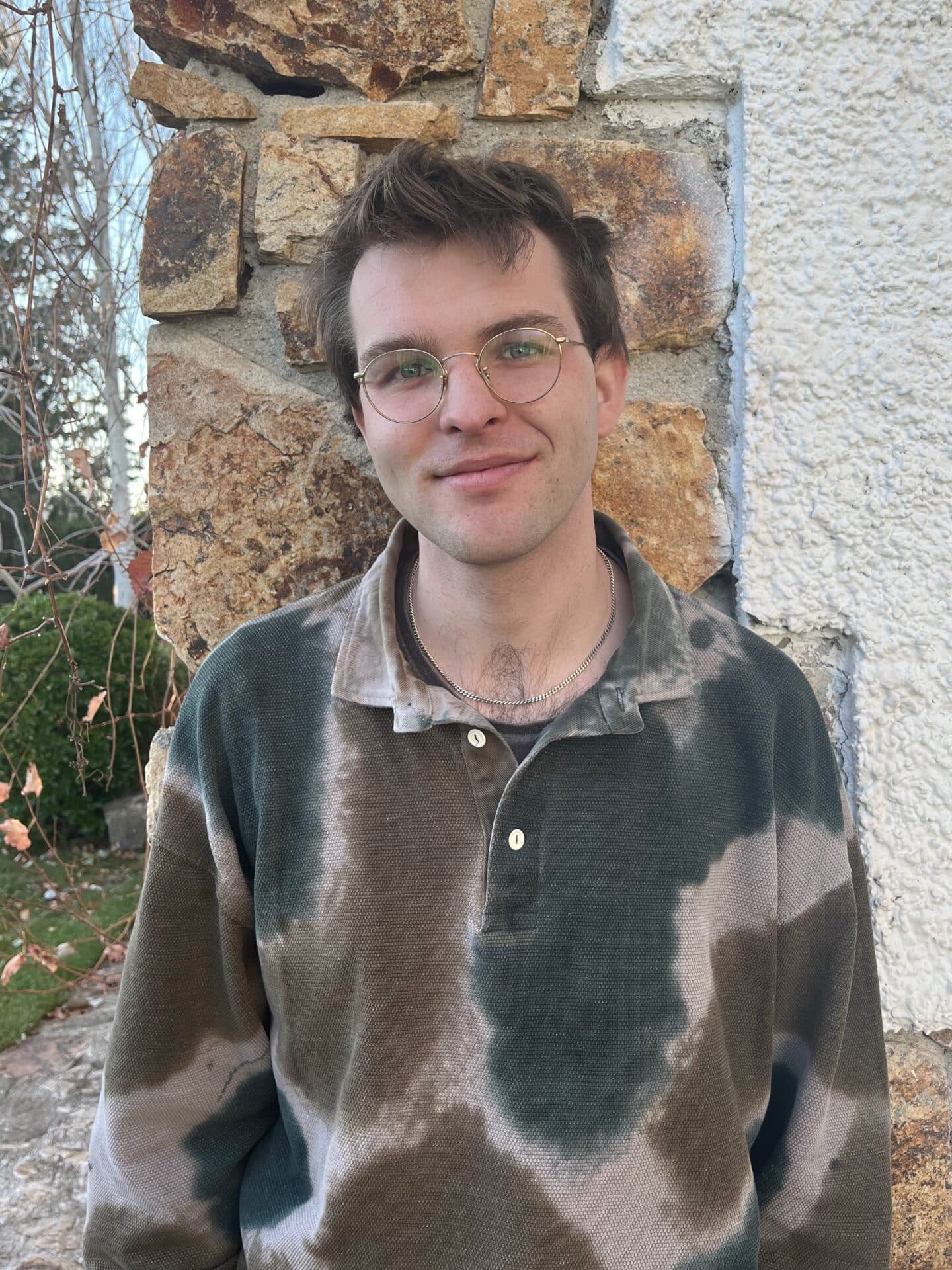 Dr Max Long standing outdoors in front of a stone and stucco wall, wearing round glasses and a tie-dye long-sleeve shirt in shades of green and beige, with trees and greenery visible in the background.