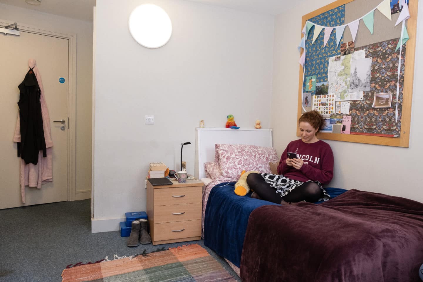 Graduate student sitting on her bed and looking at her phone in a bedroom in the EPA Centre