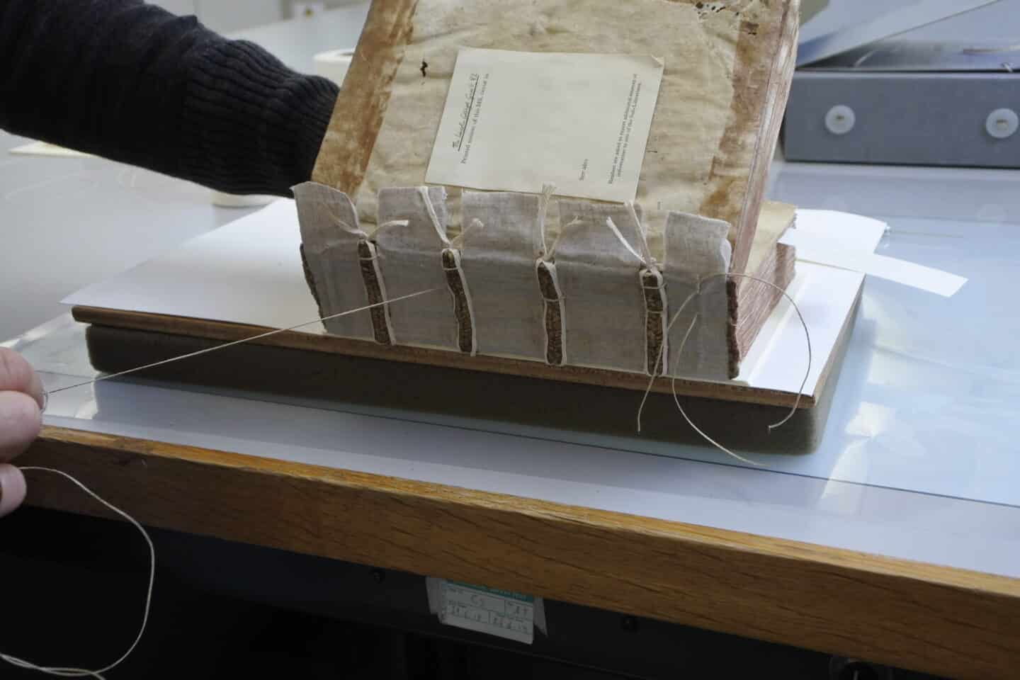 A manuscript's spine is in the process of being restored, its boards being re-attached with Japanese paper and aerolinen spine linings secured with wheat starch paste and linen braids, sewn in place with linen thread