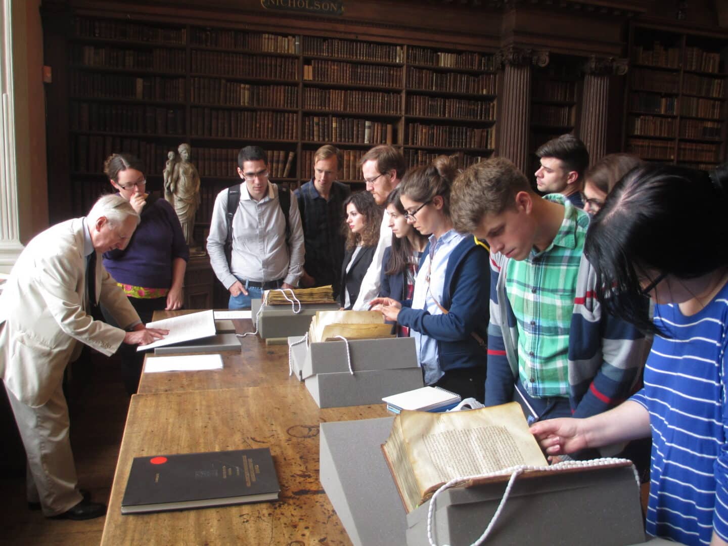 Image from a Greek summer school of a group peering at an open manuscript being explained by a man in a white suit