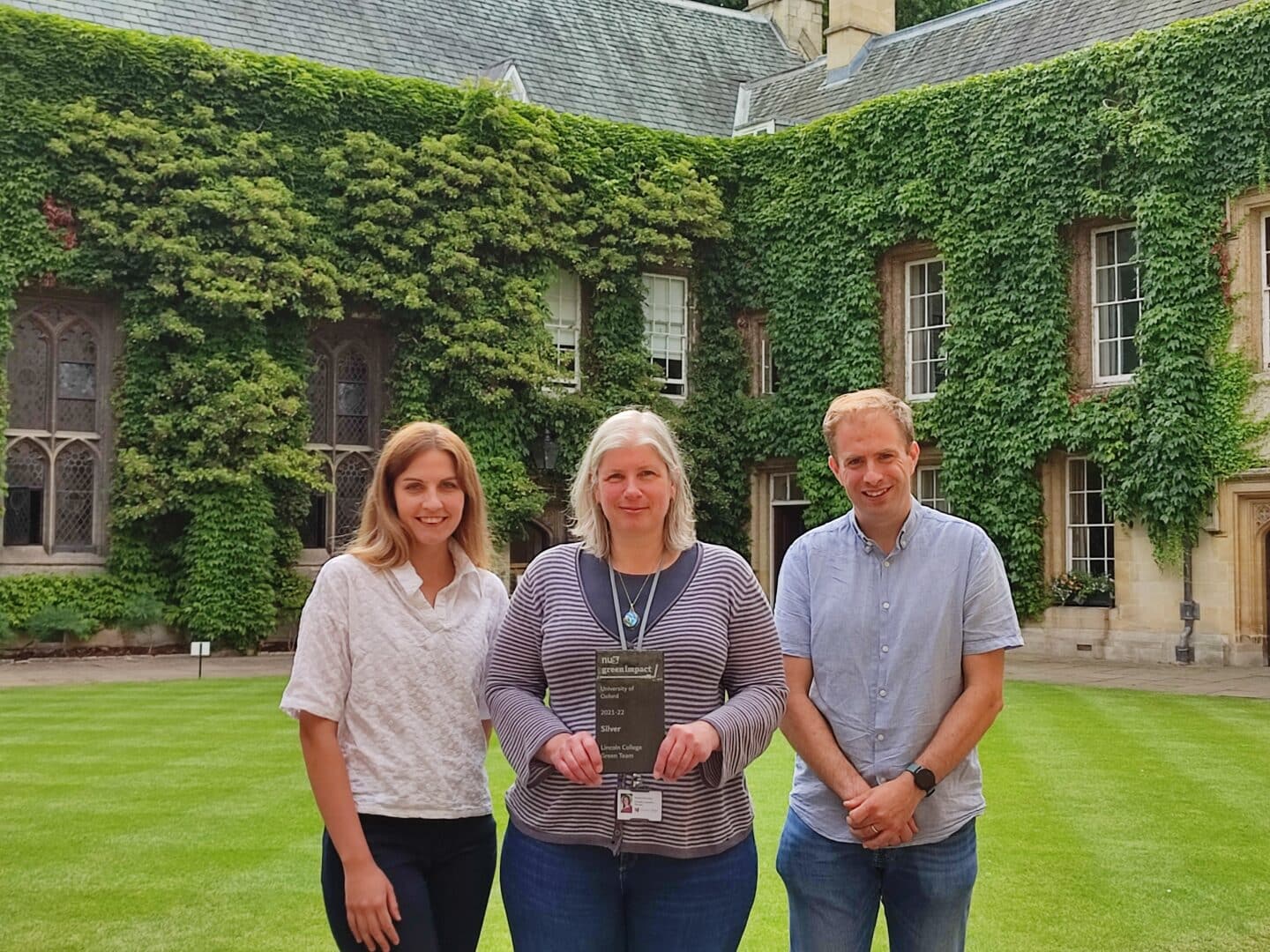 A woman, Michelle McCartney, holding a plaque awarding the Lincoln College Green Team a Silver award for green impact, flanked by a woman, Julia Uwins, and a man, Dominic Vella, all standing in a beautiful Lincoln College quad