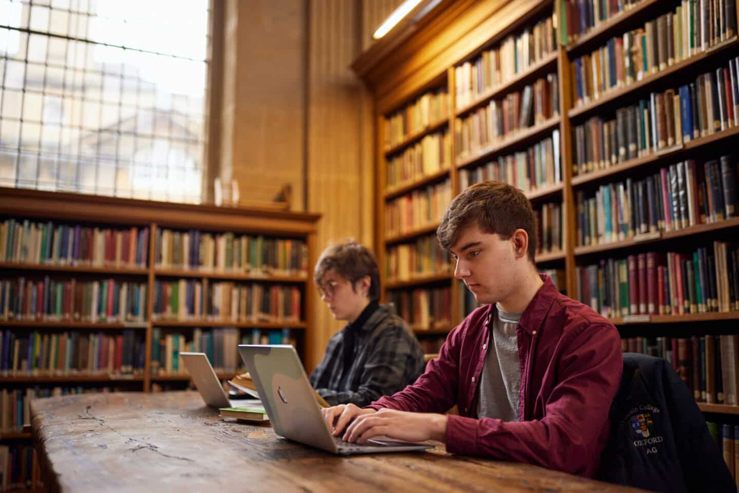 Two students studying at laptops in the Lincoln College Library, with full bookshelves behind them