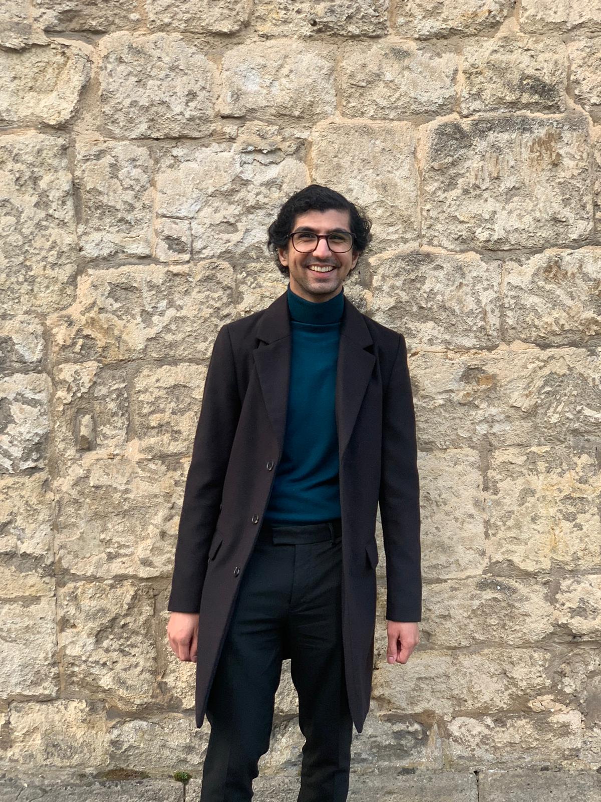 Photograph of Kishan Maher, a young man in glasses, a blue sweater and brown coat