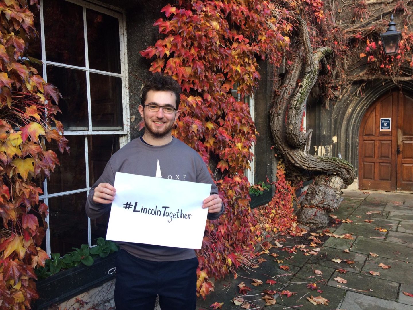 A young man standing in Lincoln College's Front Quad with red ivy in the background, holding up a sheet of paper that reads #LincolnTogether