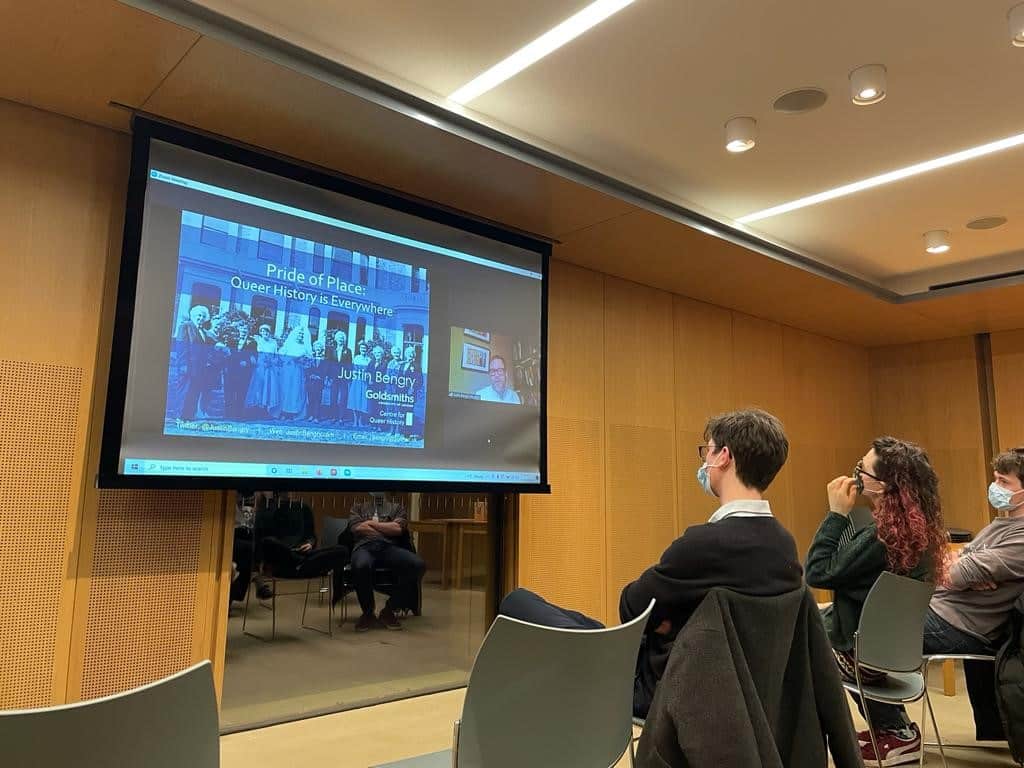 A group of students in masks sitting on chairs and watching a large screen, on which a Justin Bengry is remotely delivering a presentation entitled 'Pride of Place: Queer History is Everywhere'