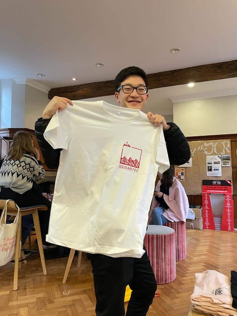 A young smiling man in glasses holding up a white t-shirt with a small logo for 'Solidaritee'