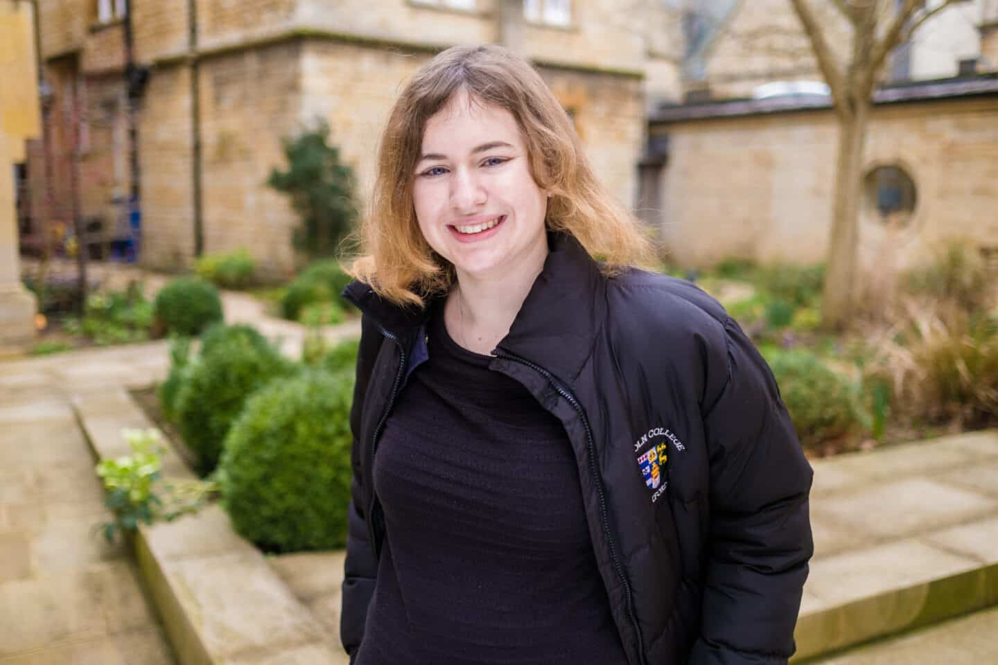 Headshot of Louise Burrett, a smiling young woman wearing a black top and a Lincoln College branded black puffer jacket