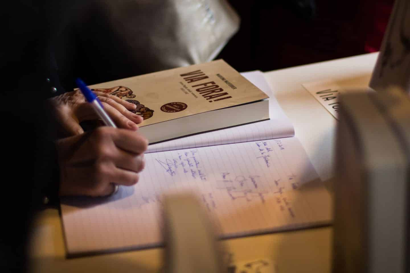 Hands making notes in a notebook with a blue pen, the book 'Via Fora!' on the side