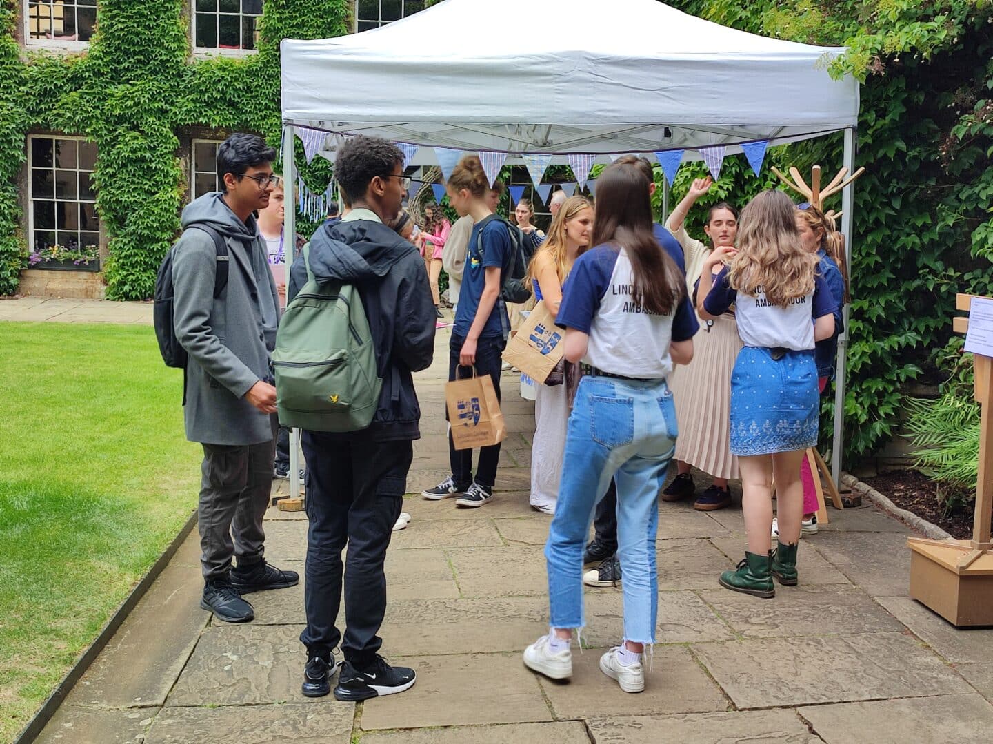 Image from a Lincoln College Open Day, featuring several Lincoln students wearing 'ambassador' t-shirts and other visitors, all under a colourful marquee in a Lincoln quad.