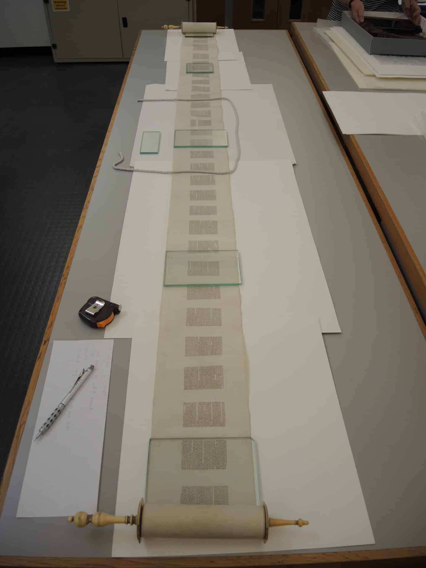 The parchment scroll is partially unrolled across a long table. A conservator looks on.