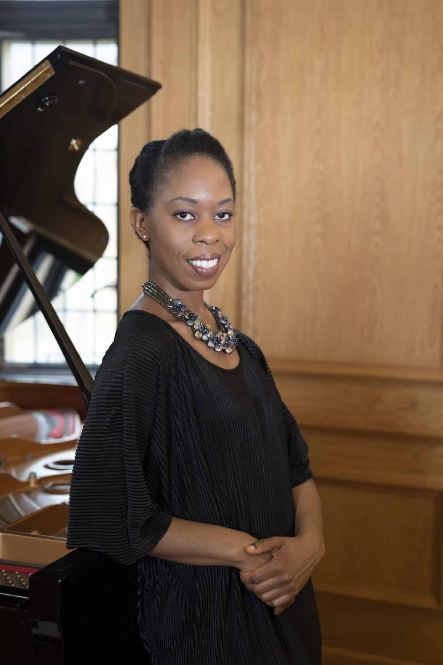 Photograph of Samantha Ege, a woman wearing a black dress and a necklace, leaning against a piano