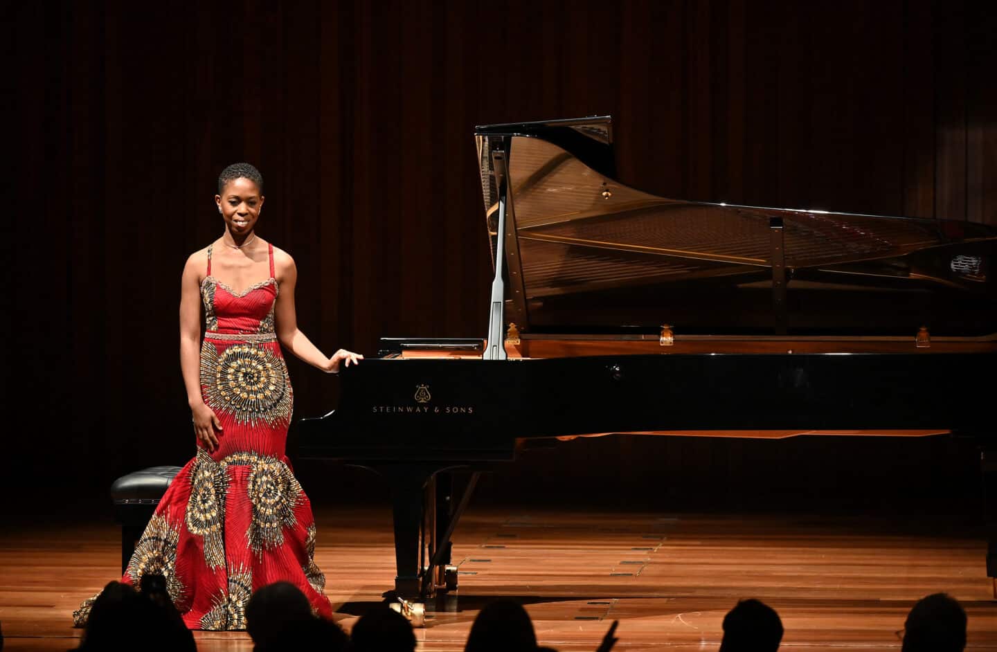 Image of Samantha Ege, a woman in a patterned red dress standing next to a piano and smiling at the audience