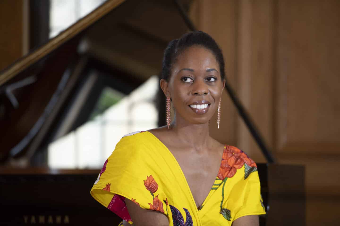 Headshot of Samantha Ege, a woman in a floral yellow dress, sitting in front of a piano that is out of focus in the background