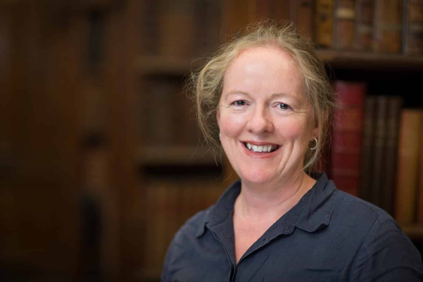 Headshot of Sarah Cusk, a woman in a black shirt with books in the background