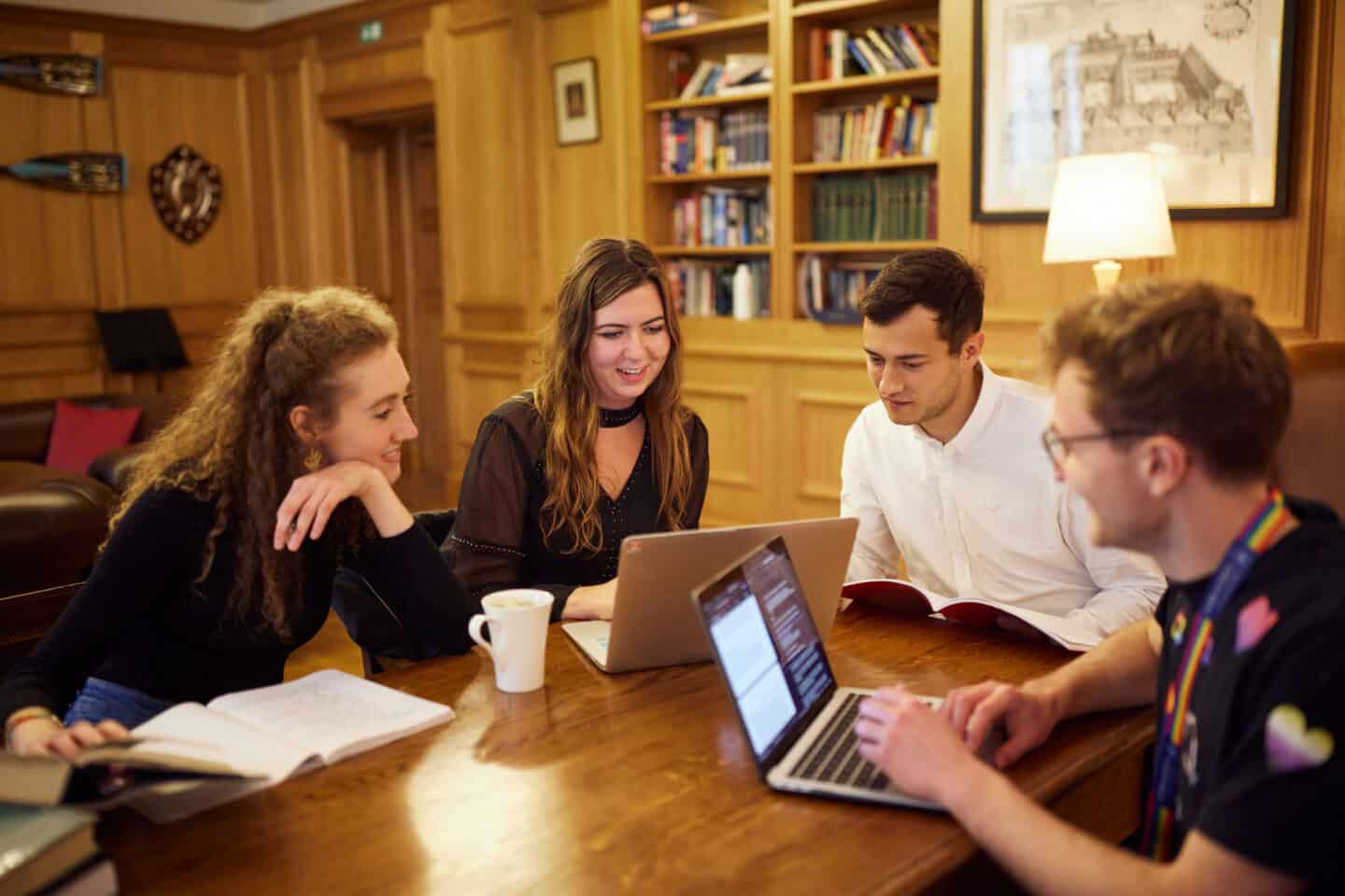 Four smiling students sitting around working with books and laptops at a wooden table in the Lincoln College Middle Common Room