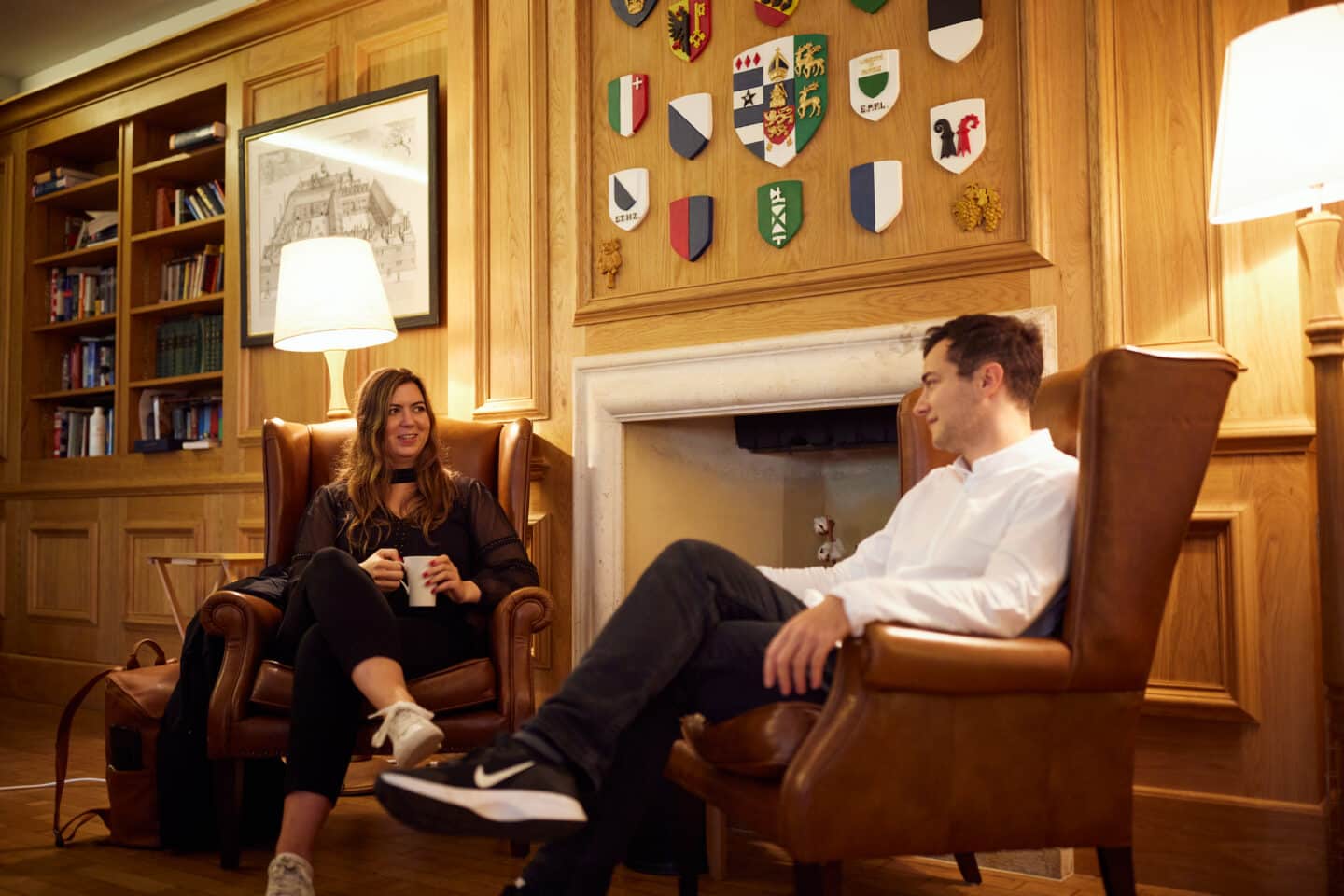 Two students sitting and chatting on large armchairs by an empty fireplace in the beautiful wood-panelled Lincoln College Middle Common Room