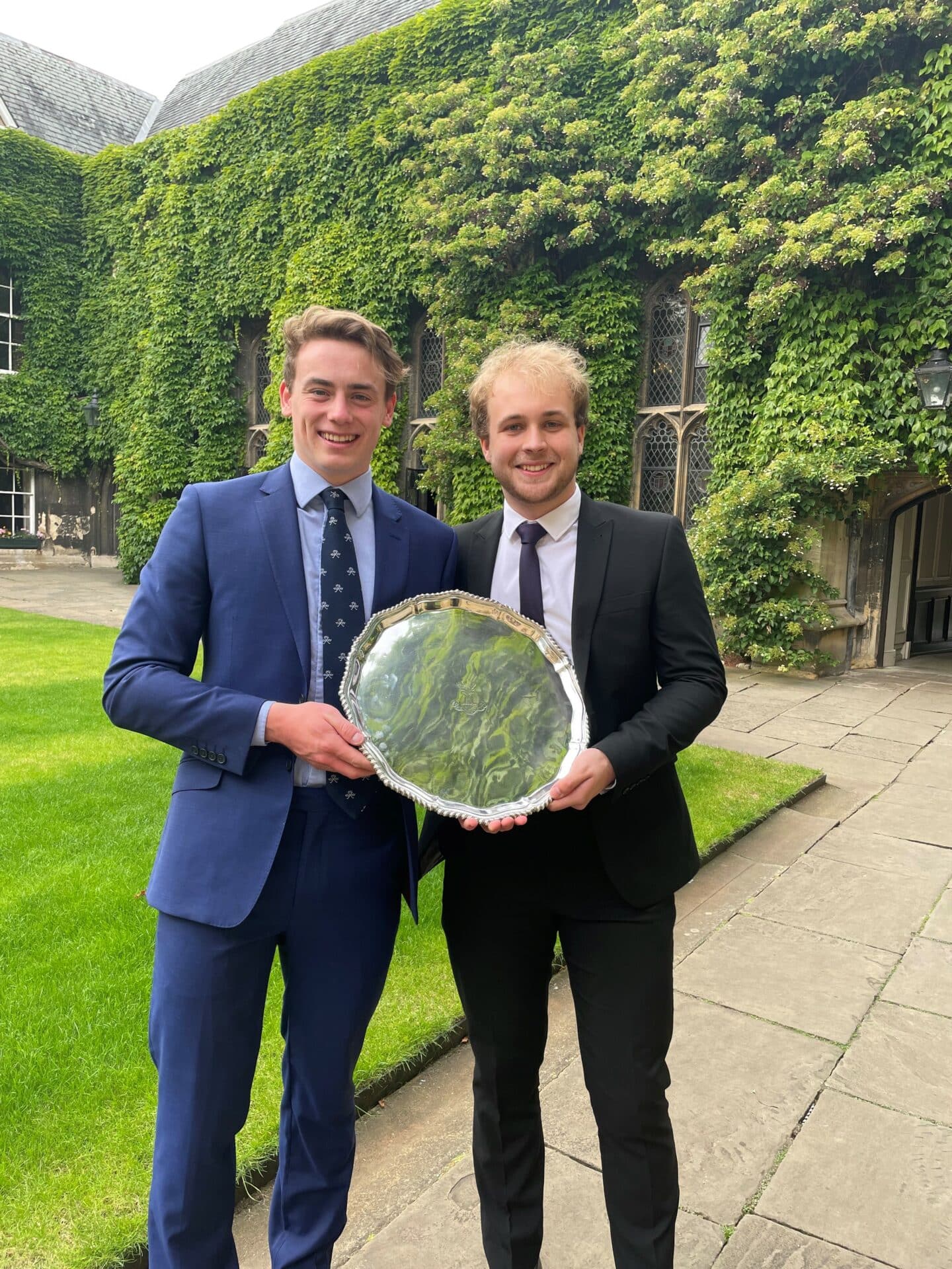 Two men in suits, smiling and holding up a trophy in the middle of a beautifully green Lincoln quad