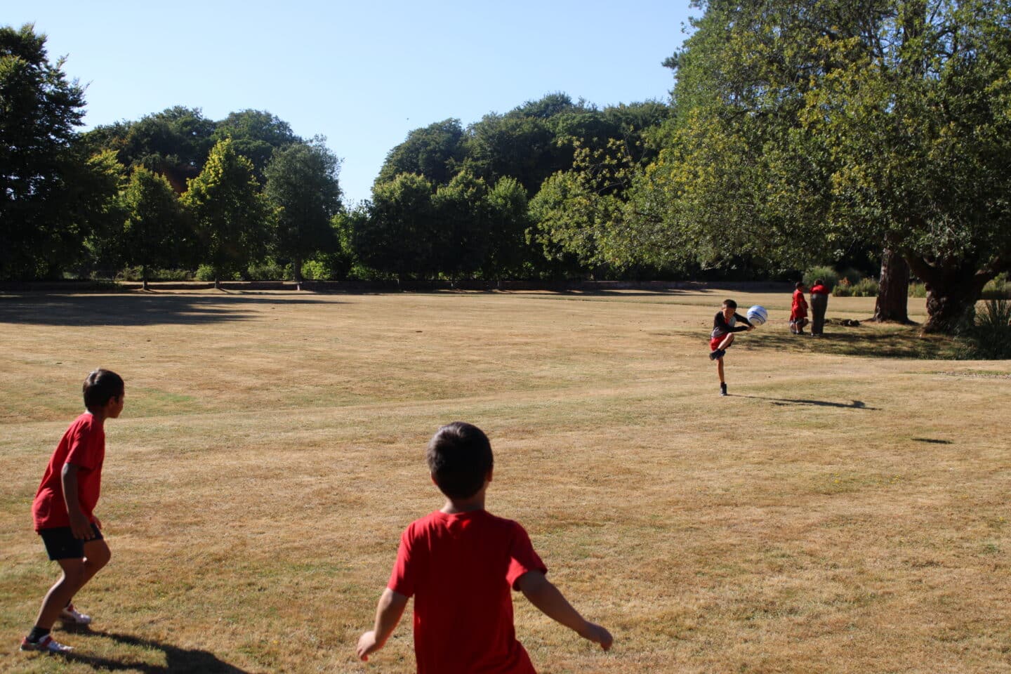 Three young boys with red t-shirts playing football on a field with trees in the background.