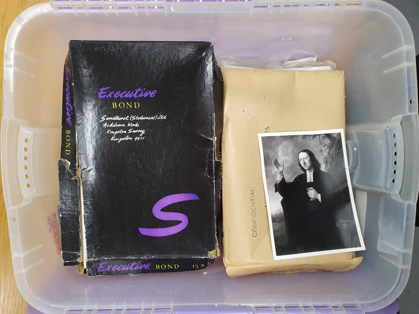 Box of items, including a black paper package labeled 'Executive Bond', a brown envelope marked 'Confidential' and a picture of a man with a long priest's collar